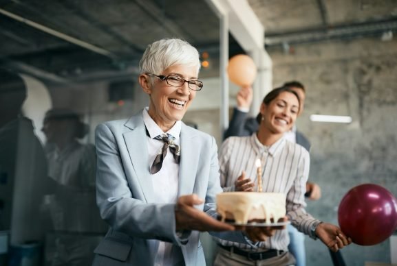 happy-senior-businesswoman-carrying-birthday-cake-on-surprise-party-in-the-office-.jpg